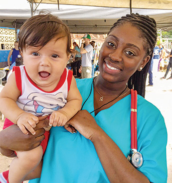 Shir 'Mel McCullough穿着蓝色的手术服，戴着红色的听诊器，抱着一个婴儿，对着镜头微笑。