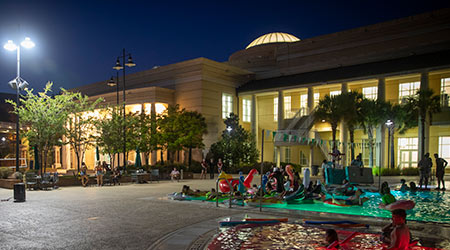 室外游泳池的夜景，周围有花车和椅子，背景是健身中心。