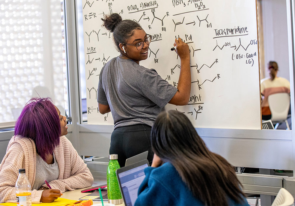 一个正在白板上写有机化学公式的学生转过身来，看着坐在桌旁的另外两个学生。