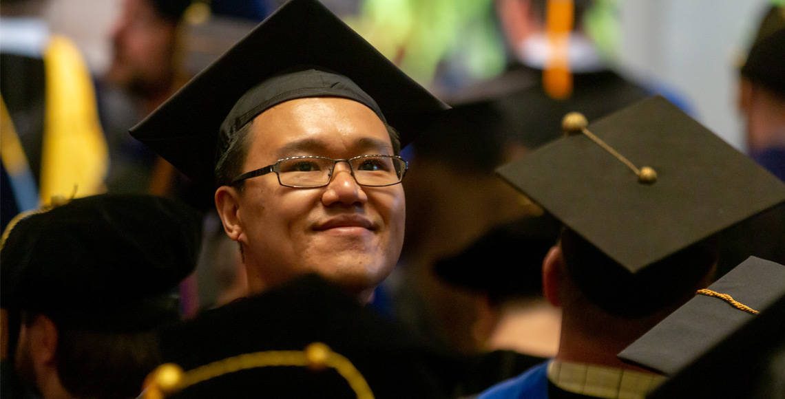 戴着毕业帽的研究生对着镜头微笑，而其他戴着毕业帽的学生则背对着镜头站在他周围。