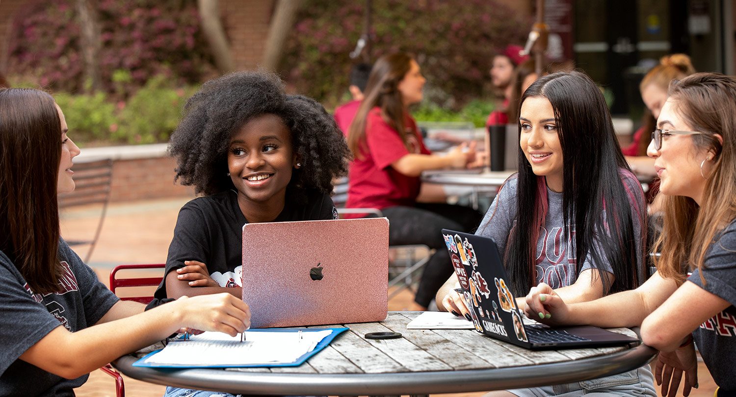 学生们聚集在外面一张桌子旁，拿着笔记本电脑学习。