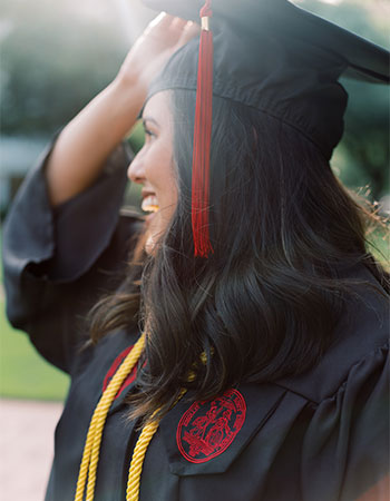 学生穿毕业帽和礼服调整她的帽子在阳光下微笑。